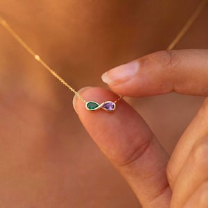 Könnte beinhalten: Goldfarbene Halskette mit einem Unendlichkeitszeichen-Anhänger. Der Anhänger hat zwei tropfenförmige Edelsteine, einen grünen und einen violetten. Die Kette wird von einem Finger gehalten.