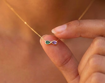 May include: A gold-colored necklace featuring an infinity symbol pendant. The pendant has two teardrop-shaped gemstones, one green and one purple. The necklace is being held by a person's finger.