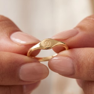 May include: A gold signet ring with a rounded rectangular face. The ring features a cursive letter "D" engraved on the face. The ring is held between two fingers, showcasing its polished finish and elegant design.