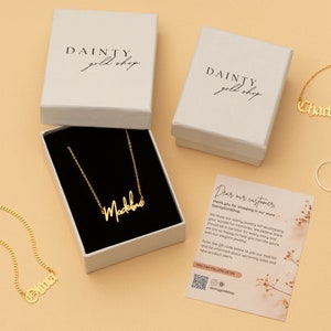 May include: Three gold necklaces with personalized names, "Olivia", "Charlotte", and "Madeline", are displayed on a light brown surface. The necklaces are packaged in white boxes with the logo "DAINTY gold shop".