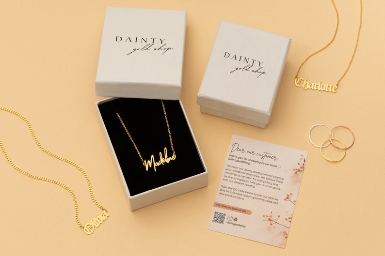 May include: Three gold necklaces with personalized names, "Olivia", "Charlotte", and "Madeline", are displayed on a light brown surface. The necklaces are packaged in white boxes with the logo "DAINTY gold shop".