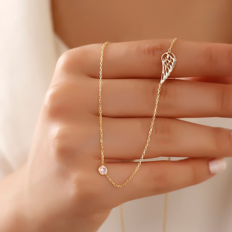 May include: A delicate gold necklace featuring a small, pink gemstone and a wing-shaped pendant. The necklace is held by a hand, showcasing the fine chain and the intricate design of the wing. The jewelry is a subtle and elegant accessory.