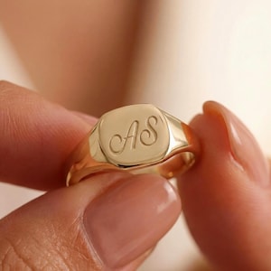 Può includere: Un anello con sigillo dorato con le iniziali "AS" incise in corsivo. L'anello ha una faccia quadrata e una finitura lucida. L'anello è tenuto tra le dita, mettendo in risalto il suo design e il suo colore.