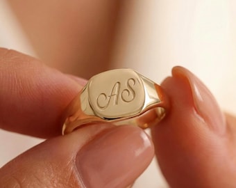 May include: A gold signet ring featuring the engraved initials "AS" in a cursive style. The ring has a square face and a shiny finish. The ring is held between fingers, highlighting its design and colour.