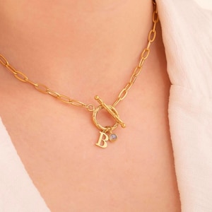 May include: A gold chain necklace with a toggle clasp and a charm featuring the letter "B" and a small gemstone.