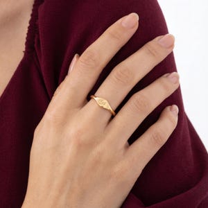 Puede incluir: Un anillo de sello de oro con una superficie lisa y pulida que presenta una pequeña letra D grabada en cursiva en el centro de la cara ovalada. El anillo tiene una banda fina y redondeada con un acabado amarillo metálico cálido y brillante.