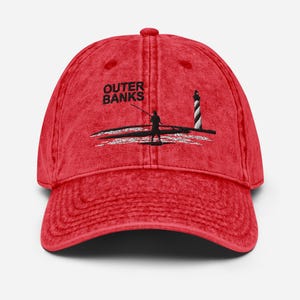 May include: Red baseball cap with the text "Outer Banks" above a black and white illustration of a lighthouse and a person fishing. The cap has a curved brim and a vintage, washed look.