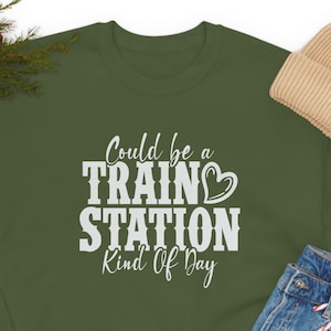 May include: An olive green sweatshirt with the text "Could be a TRAIN STATION Kind Of Day" in white. The image also includes a beige beanie, blue jeans, and black trainers.