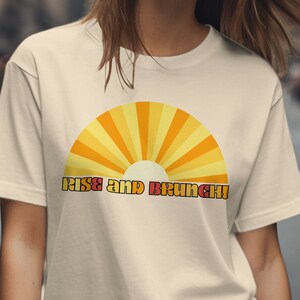 May include: A beige t-shirt with a retro-style sunrise graphic and the text "Rise and Brunch!" printed in red and orange.