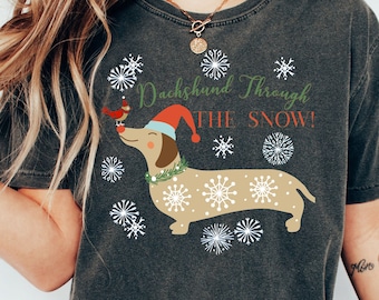 May include: A dark grey t-shirt featuring a cartoon dachshund wearing a Santa hat and carrying a bird. The text on the shirt reads "Dachshund Through The Snow!" with snowflakes surrounding the dachshund.