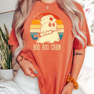 May include: A red t-shirt with a retro-style graphic of a ghost wearing a nurse's hat and holding a syringe. The text "BOO BOO CREW" is printed below the graphic.