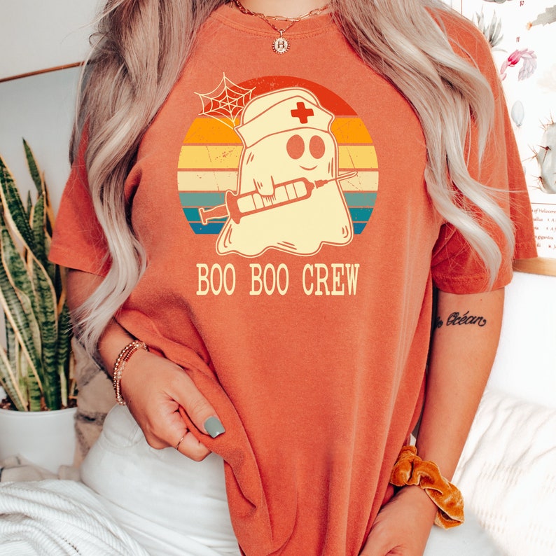 May include: A red t-shirt with a retro-style graphic of a ghost wearing a nurse's hat and holding a syringe. The text "BOO BOO CREW" is printed below the graphic.