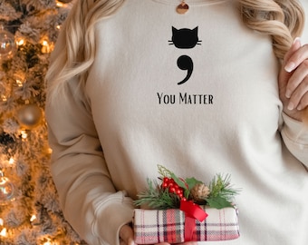 May include: A beige sweatshirt with a black cat silhouette and a semicolon, with the text "You Matter" below. The sweatshirt is being held by a person in front of a Christmas tree.