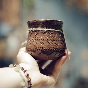 May include: A small, handcrafted brown ceramic pot with etched geometric patterns. The pot is wrapped with twine and held in a hand, suggesting a rustic or artisanal aesthetic. The pot is likely used for decorative or functional purposes.