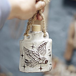 May include: White ceramic bottle featuring an owl design and star details. It has a cork stopper and a woven rope handle. A decorative item for the home, ideal for displaying.