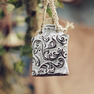 Può includere: Piccola bottiglia in ceramica bianca con tappo in sughero e manico in corda di iuta. La bottiglia è decorata con un motivo floreale a volute in rilievo, marrone scuro. Lo sfondo è sfocato, con accenni di fogliame verde.