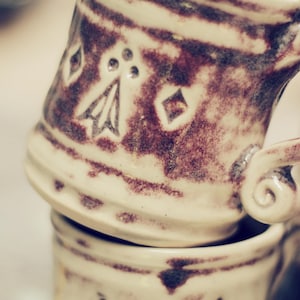May include: A stack of ceramic mugs featuring a cream-colored glaze with dark brown, mottled patches. Each mug is decorated with embossed geometric diamond shapes and a stylized heraldic ermine spot pattern, and includes a small, curled handle.
