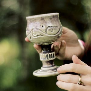Pode incluir: Um cálice de cerâmica com esmalte esbranquiçado salpicado e detalhes em castanho escuro. A taça apresenta um padrão esculpido em relevo com volutas, enquanto o pé é estruturado com estrias verticais que descem até uma base circular sombreada.