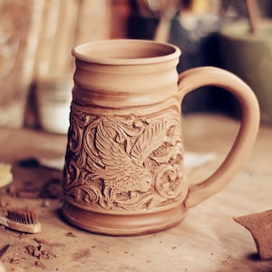 Puede incluir: Una taza de cerámica marrón con un diseño detallado e intrincado de un pájaro y motivos florales. La taza tiene un asa y está hecha a mano.