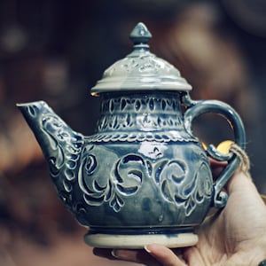 May include: A blue ceramic teapot with a lid, spout, and handle. The teapot features intricate floral and scrollwork designs. The handle is arched, and the spout is curved. The teapot is held by a hand.