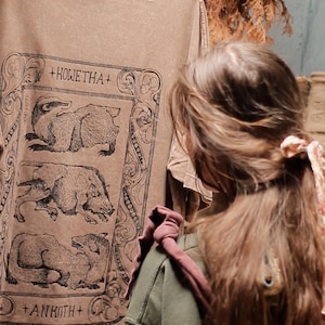 May include: A brown t-shirt featuring a black ink graphic print of three stacked animals within an ornate rectangular frame. The text +KOWETHA+ appears above the top animal, and +ANKOTH+ is printed below the bottom animal in a serif font.