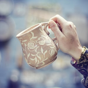 May include: A beige ceramic mug with a handle, decorated with a white pomegranate and leaf design. The mug is held in a hand, highlighting the floral pattern. A unique piece of pottery.