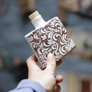 Può includere: Una bottiglia in ceramica marrone e bianca con un tappo di sughero. La bottiglia ha una forma quadrata con un motivo floreale bianco su sfondo marrone. La bottiglia è tenuta in mano da una persona.