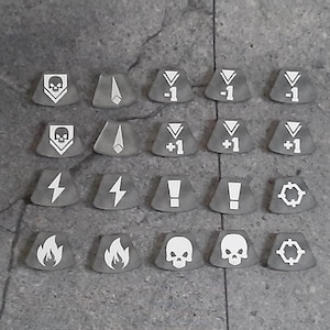 May include: A set of 18 grey tokens with various white symbols, including skulls, lightning bolts, flames, and numbers. The tokens are arranged in a grid pattern on a grey surface.