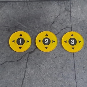 Könnte beinhalten: Drei gelbe runde Spielmarken mit schwarzen Zahlen 1, 2 und 3 sowie Richtungspfeilen. Die Spielmarken sind in einer horizontalen Linie auf einer grauen, strukturierten Oberfläche angeordnet.