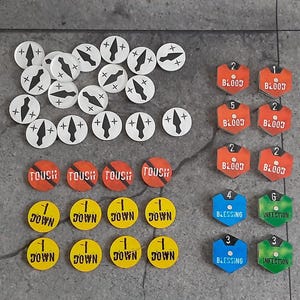 Puede incluir: Una colección de fichas de juego de varias formas y colores. Hay fichas circulares blancas con diseños de flechas negras, fichas en forma de escudo naranja con la palabra "BLOOD" y fichas circulares amarillas con la palabra "DOWN". Otras fichas son azules y verdes.