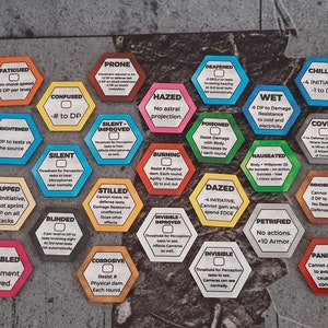 May include: A set of 20 hexagonal game tokens with various colors and text descriptions. The tokens are made of cardboard and have a glossy finish. The text on the tokens describes different conditions or effects that can be applied to players in a game.
