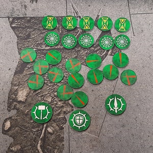 Op de afbeelding: Een verzameling groene, cirkelvormige spel fiches met diverse witte en oranje symbolen. De fiches zijn gerangschikt op een grijze ondergrond. De symbolen omvatten zandlopers, kruisen en andere geometrische ontwerpen. De fiches lijken van hout of een vergelijkbaar materiaal te zijn.