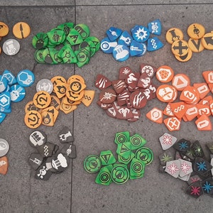 Puede incluir: Una colección de fichas de juego coloridas con varios símbolos y diseños. Las fichas son de plástico y están dispuestas en filas sobre una superficie gris.