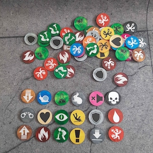 Puede incluir: Una colección de fichas de juego circulares y coloridas con varios símbolos. Las fichas presentan iconos como escudos, llamas y conejos, en colores como verde, rojo y amarillo. Las fichas están dispersas sobre una superficie gris.