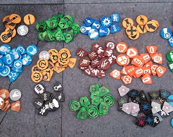May include: A collection of assorted game tokens in various shapes and colors, including orange, green, blue, brown, black, and white. The tokens feature diverse printed icons such as arrows, skulls, gears, and geometric symbols for tabletop gaming.