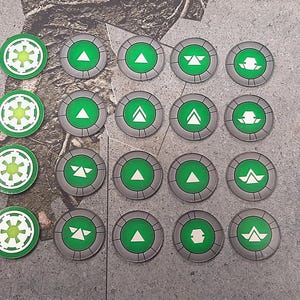 Puede incluir: Una colección de fichas de juego circulares. Algunas fichas son verdes con símbolos blancos, mientras que otras son grises con centros verdes y símbolos blancos. Las fichas están dispuestas sobre una superficie texturizada.