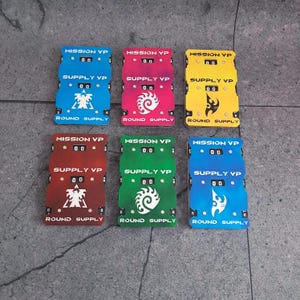 May include: Six rectangular game score trackers in blue, pink, yellow, red, green, and light blue. Each tracker features three sections labelled MISSION VP, SUPPLY VP, and ROUND SUPPLY with mechanical number dials set to 00 and unique white icons.