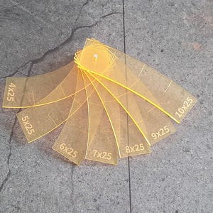 May include: A set of clear, yellow acrylic quilting templates, each labeled with measurements in inches. The templates are arranged in a fan shape, with curved edges and a central hole for storage. The background is a textured gray surface.