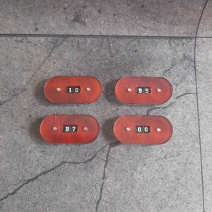 May include: Four oval-shaped, reddish-brown translucent plastic counters, each featuring a central mechanical display window showing two-digit numbers. The counters display the numbers 10, 99, 87, and 00, with small silver-coloured rivets on each side.