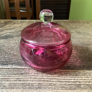 May include: A round, translucent pink glass jar with a matching lid. The lid features a clear, spherical glass knob handle at the top. The jar has a smooth, curved body and sits as a standalone container, showcasing a vibrant magenta hue throughout.