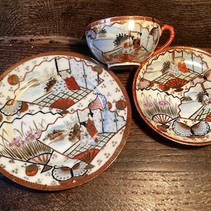 May include: A vintage Japanese teacup and saucer set with a delicate floral and geometric design. The set features a white background with intricate gold and red accents. The cup and saucer are decorated with images of women in traditional Japanese clothing.