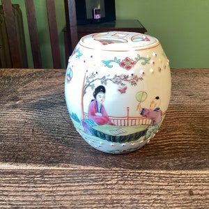 May include: A white porcelain jar with a lid, decorated with a colourful scene of two figures in a garden setting. The jar has a textured surface with small white dots.