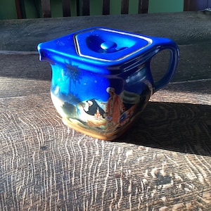 May include: A blue ceramic teapot with a lid and a handle. The teapot features a painted scene of people in a desert setting.