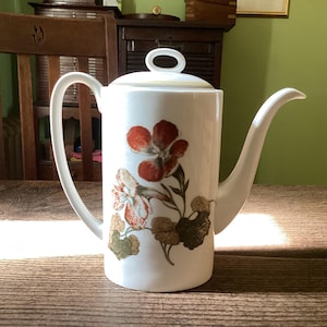 May include: White ceramic coffee pot with a floral design featuring red, orange, and green flowers. The pot has a curved spout and a handle on the side. The lid has a small loop handle. The pot is sitting on a wooden table.