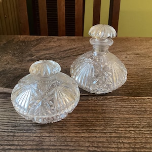 May include: Two clear glass bottles with intricate cut glass designs. The bottles have a round shape and a decorative stopper.