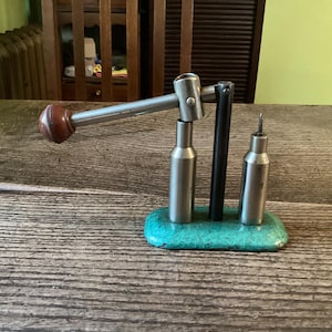 May include: A metal reloading tool with a wooden handle and a teal base. The tool has a lever, two cylindrical components, and a pointed component. The tool is designed for reloading ammunition.