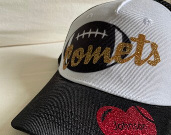 Può includere: Un cappello da baseball bianco e nero con una grafica di football bianco e nero e la parola "Comets" in glitter dorato. Il cappello ha una parte posteriore in rete nera e un cuore rosso con la parola "Johnson" in glitter bianco.