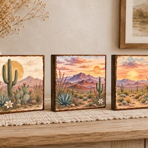May include: Three square wooden wall art panels featuring desert landscapes. Each panel displays a sunset scene with mountains, saguaro cacti, blooming prickly pear cacti, and agave plants in shades of orange, pink, green, and brown.