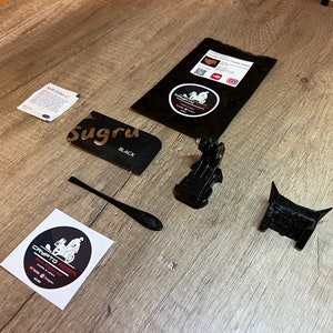 May include: A flat lay of various items for a 3D printed motorcycle mount. The items include a black Sugru adhesive, a black plastic tool, a black 3D printed motorcycle mount, a black 3D printed GoPro mount, a black package with a white sticker, and a white sticker with a black and white illustration of a motorcycle.