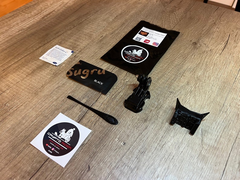 May include: A flat lay of various items for a 3D printed motorcycle mount. The items include a black Sugru adhesive, a black plastic tool, a black 3D printed motorcycle mount, a black 3D printed GoPro mount, a black package with a white sticker, and a white sticker with a black and white illustration of a motorcycle.